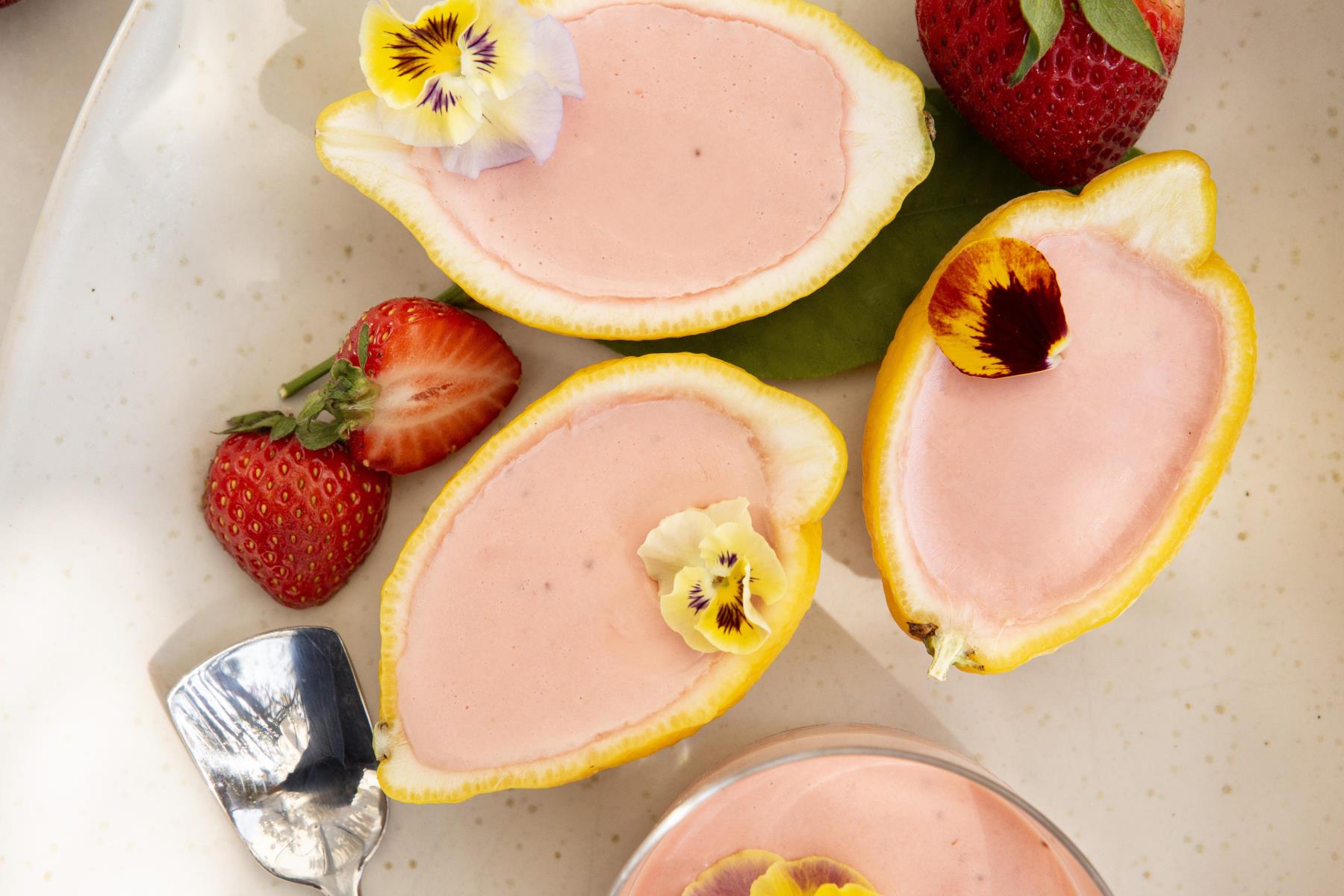 Strawberry posset served in lemon halves, garnished with edible flowers, and accompanied by fresh strawberries on a white plate with a silver spoon.