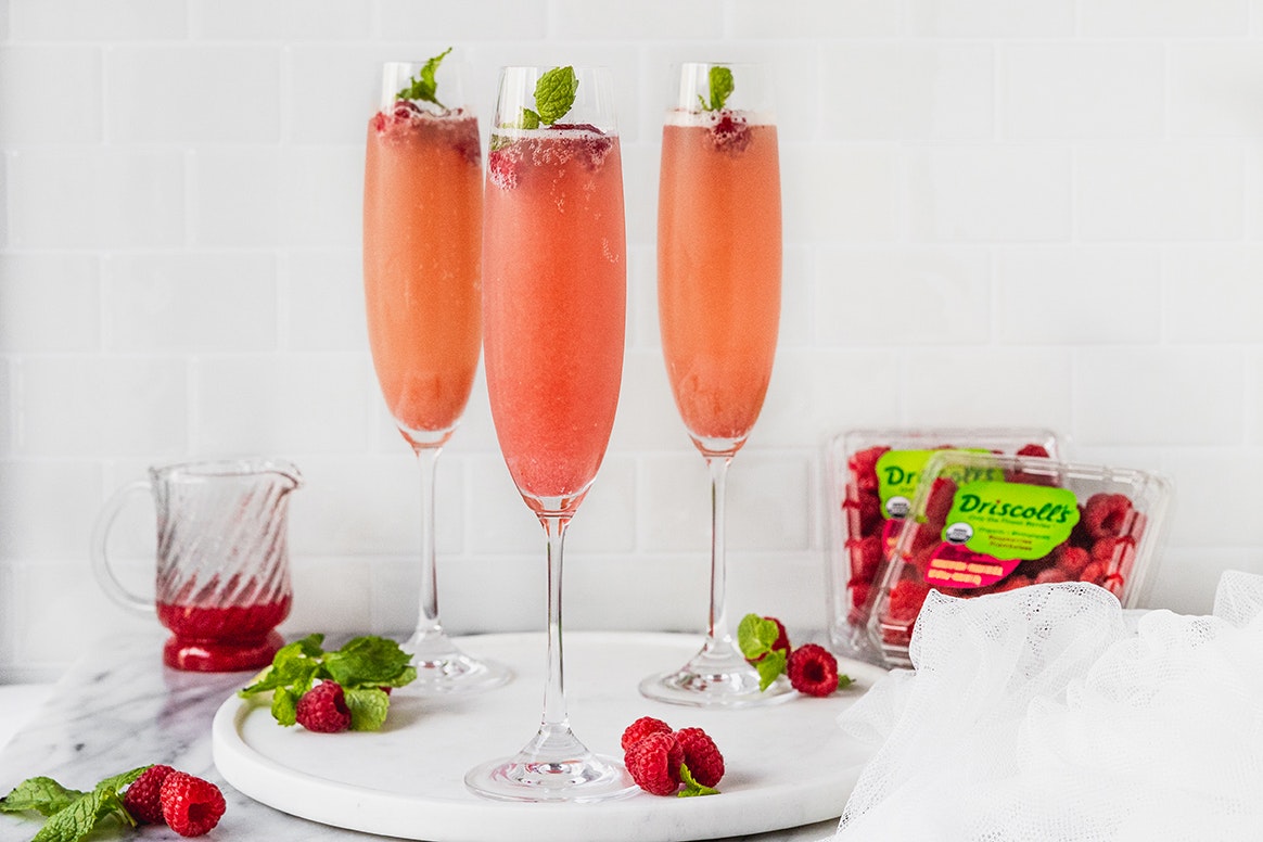 Raspberry mimosas topped with fresh raspberries and garnish. Driscoll's clamshells of organic raspberries along with a small container filled with raspberry sauce sit on the counter behind the champagne glasses. The raspberry mimosas are sitting on a white plate with raspberries and garnish scattered throughout the photo.