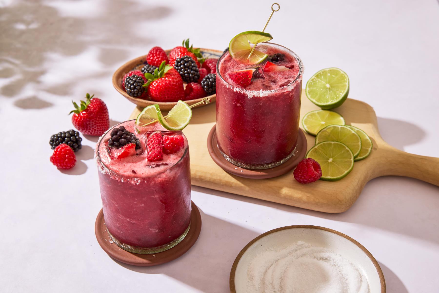 Two berry margaritas garnished with lime and fresh berries, alongside a bowl of mixed berries and lime slices.